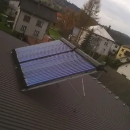 Kolektory słoneczne zamontowane na metalowym dachu domu jednorodzinnego, widok z góry na tle zabudowy mieszkalnej i zalesionych wzgórz w pochmurny dzień.