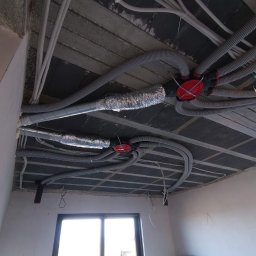 HYDRO-HOUS - Instalacja przewodów wentylacyjnych i elektrycznych pod betonowym stropem w surowym wnętrzu, widoczne przewody elastyczne, rury aluminiowe i rozdzielacze z czerwonymi pokrywami.