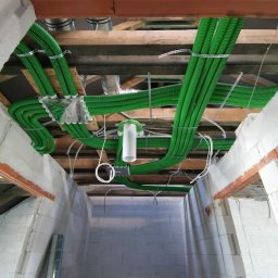 HYDRO-HOUS - Widok z dołu na zielone, karbowane rury wentylacyjne zamontowane pod drewnianym stropem w surowym wnętrzu budynku z białych bloczków.