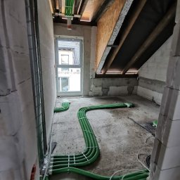 HYDRO-HOUS - Wnętrze w stanie surowym z widoczną instalacją wentylacyjną w postaci zielonych rur rozprowadzonych po podłodze i podwieszonych pod sufitem, zabezpieczonych białymi opaskami zaciskowymi.