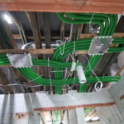 HYDRO-HOUS - Sufit poddasza z widocznymi zielonymi rurami wentylacyjnymi przymocowanymi do drewnianej konstrukcji, białe skrzynki rozdzielcze i fragment surowej ściany z białych bloczków.