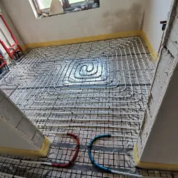 Układ rur grzewczych na podłodze, przygotowany do zalania wylewką, widok z góry, rury ułożone spiralnie na folii izolacyjnej, fragment ściany w surowym stanie.