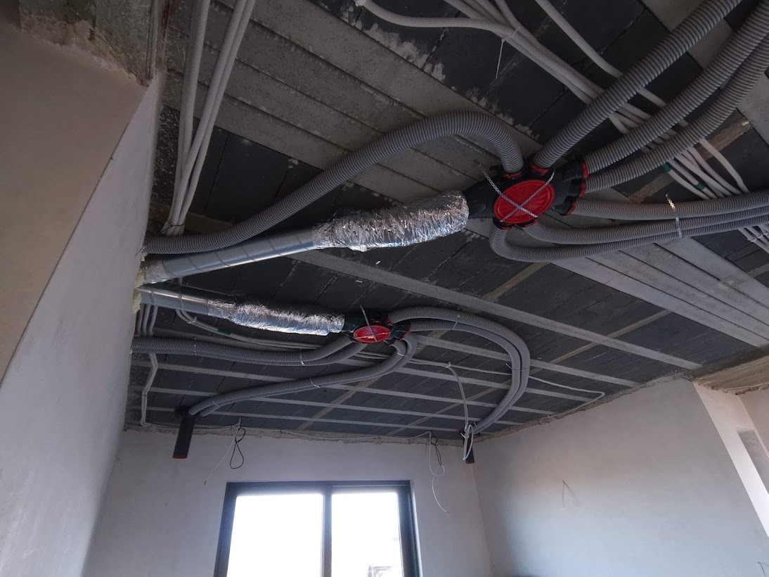 Instalacja przewodów wentylacyjnych i elektrycznych pod betonowym stropem w surowym wnętrzu, widoczne przewody elastyczne, rury aluminiowe i rozdzielacze z czerwonymi pokrywami.