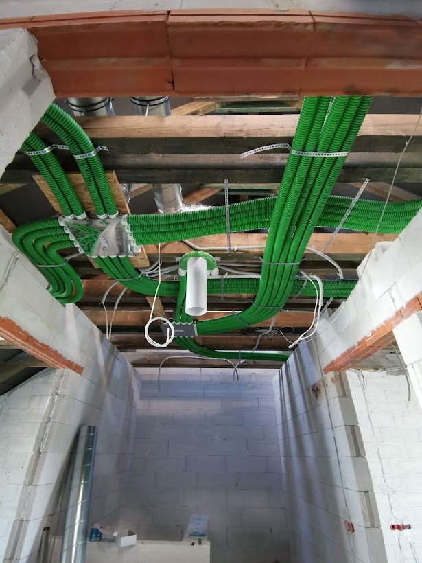 Widok z dołu na zielone, karbowane rury wentylacyjne zamontowane pod drewnianym stropem w surowym wnętrzu budynku z białych bloczków.