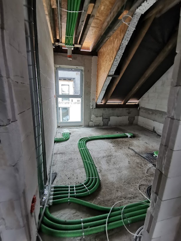 Wnętrze w stanie surowym z widoczną instalacją wentylacyjną w postaci zielonych rur rozprowadzonych po podłodze i podwieszonych pod sufitem, zabezpieczonych białymi opaskami zaciskowymi.