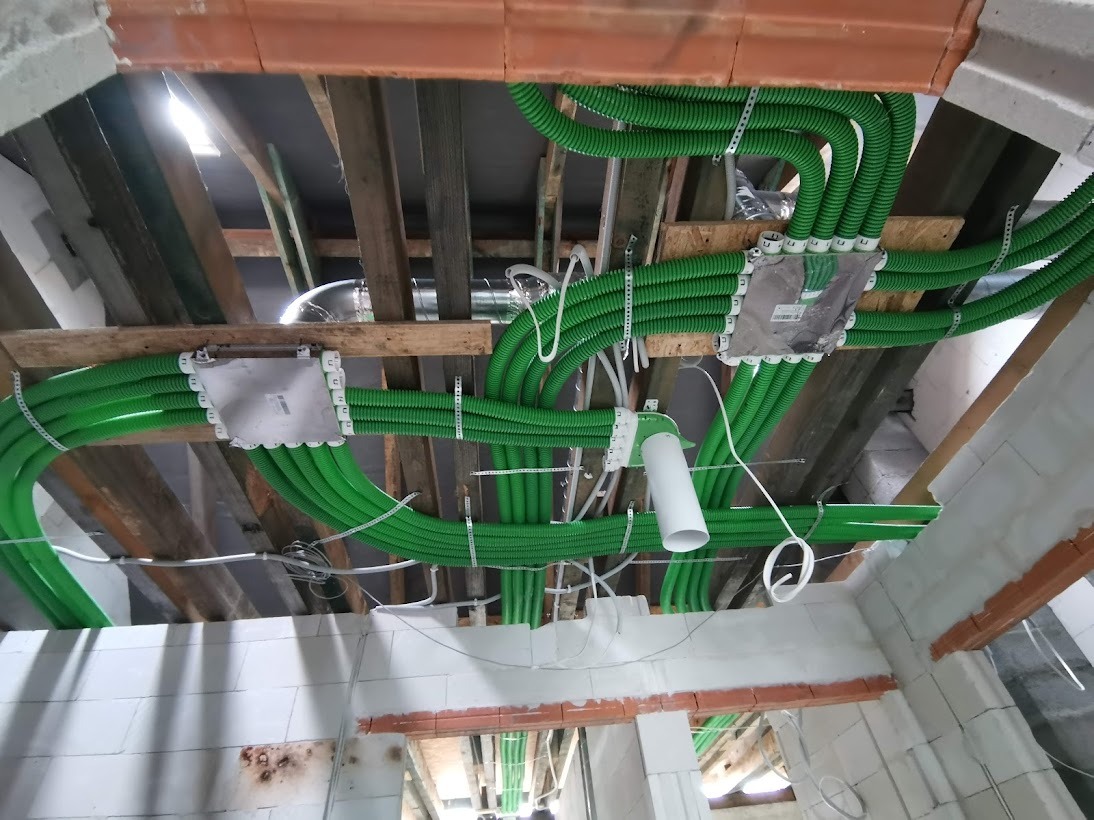 Sufit poddasza z widocznymi zielonymi rurami wentylacyjnymi przymocowanymi do drewnianej konstrukcji, białe skrzynki rozdzielcze i fragment surowej ściany z białych bloczków.