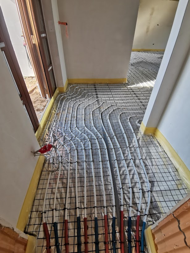 Instalacja ogrzewania podłogowego w trakcie montażu: białe rurki ułożone na czarnej siatce z folią odblaskową, widoczne wystające rury w kolorze czerwonym i niebieskim, żółta taśma przy ścianach.