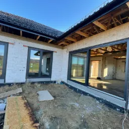 Dom w trakcie budowy, widoczne czarne ramy okienne w surowych ścianach z betonu komórkowego, dach pokryty dachówką, teren wokół domu jeszcze niezagospodarowany.