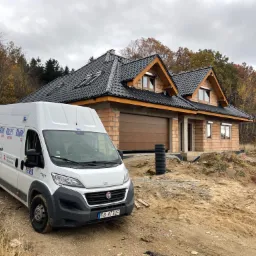 Nowy dom w trakcie budowy z zamontowanymi oknami i brązową bramą garażową, na pierwszym planie zaparkowany biały bus firmy budowlanej z reklamą okien, drzwi, rolet i bram.