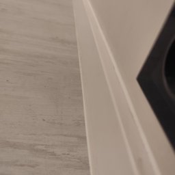 Remontowo - Szczegół wykończenia wnętrza: połączenie podłogi imitującej drewno i pomalowanej na biało ściany z widocznym fragmentem otworu wentylacyjnego.