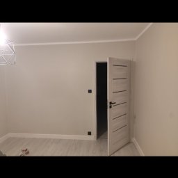 Remontowo - Jasne, świeżo wykończone wnętrze pokoju z otwartymi, drewnianymi drzwiami z poziomymi frezami, szarymi ścianami i panelami podłogowymi, widoczna nowoczesna lampa wisząca.