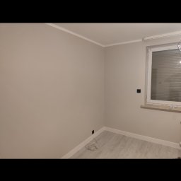 Remontowo - Pomalowane na beżowo ściany pokoju z szarą podłogą, białą listwą przypodłogową i sufitową oraz oknem z białymi żaluzjami.