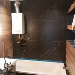 Remontowo - Nowoczesna łazienka z czarnymi płytkami imitującymi marmur, drewnianymi panelami, białą wanną i podgrzewaczem wody Junkers na ścianie.