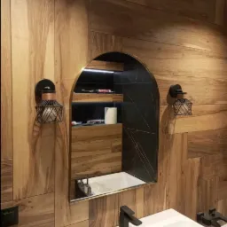 Nowoczesna łazienka z drewnopodobnymi płytkami na ścianach, białą umywalką z czarną armaturą, lustrem w kształcie łuku z czarnym obramowaniem i wnęką z oświetleniem LED, gdzie widoczne są akcesoria...