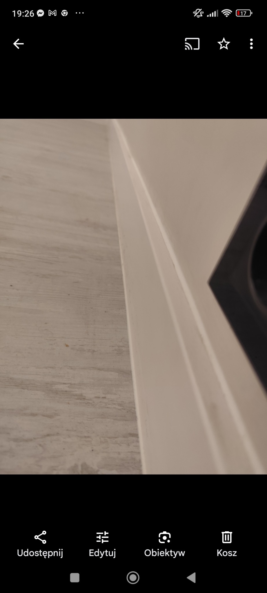 Szczegół wykończenia wnętrza: połączenie podłogi imitującej drewno i pomalowanej na biało ściany z widocznym fragmentem otworu wentylacyjnego.