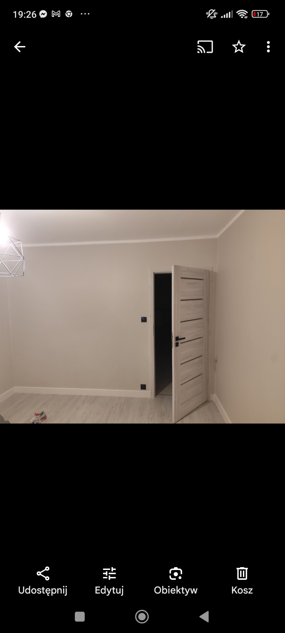 Jasne, świeżo wykończone wnętrze pokoju z otwartymi, drewnianymi drzwiami z poziomymi frezami, szarymi ścianami i panelami podłogowymi, widoczna nowoczesna lampa wisząca.