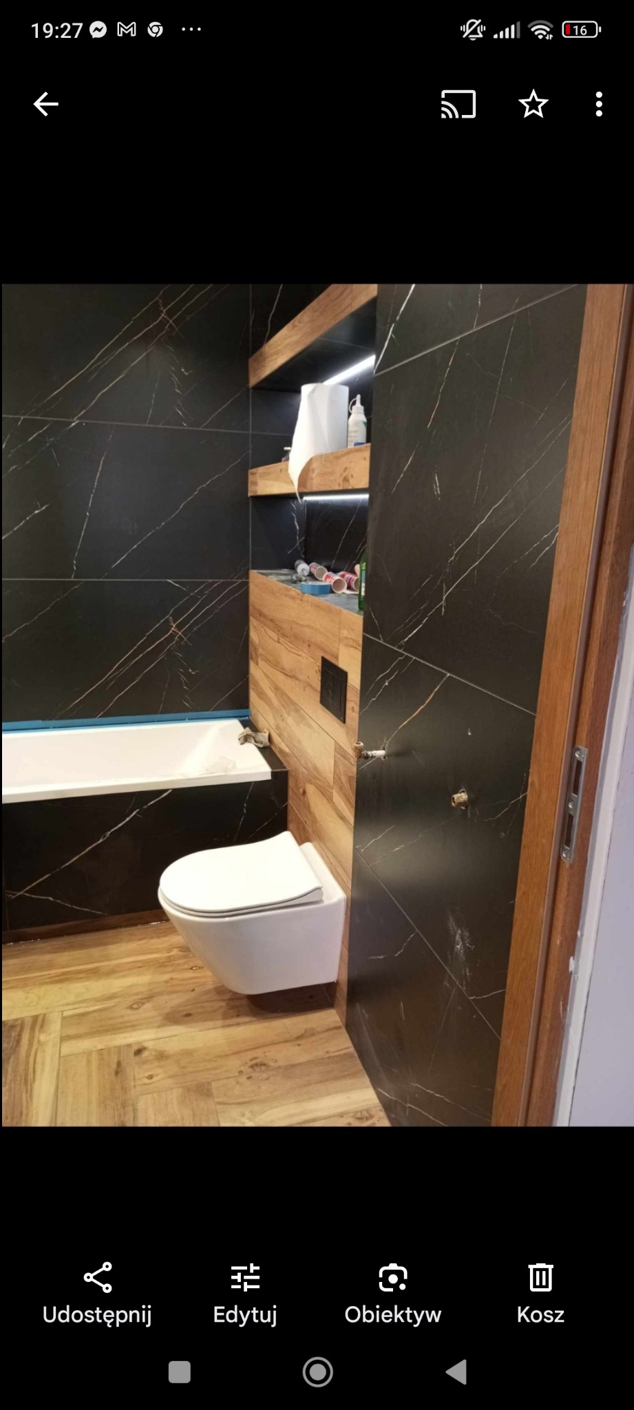 Nowoczesna łazienka z czarnymi płytkami imitującymi marmur, drewnianymi akcentami i podwieszaną toaletą. Na półkach widoczne kosmetyki i akcesoria.