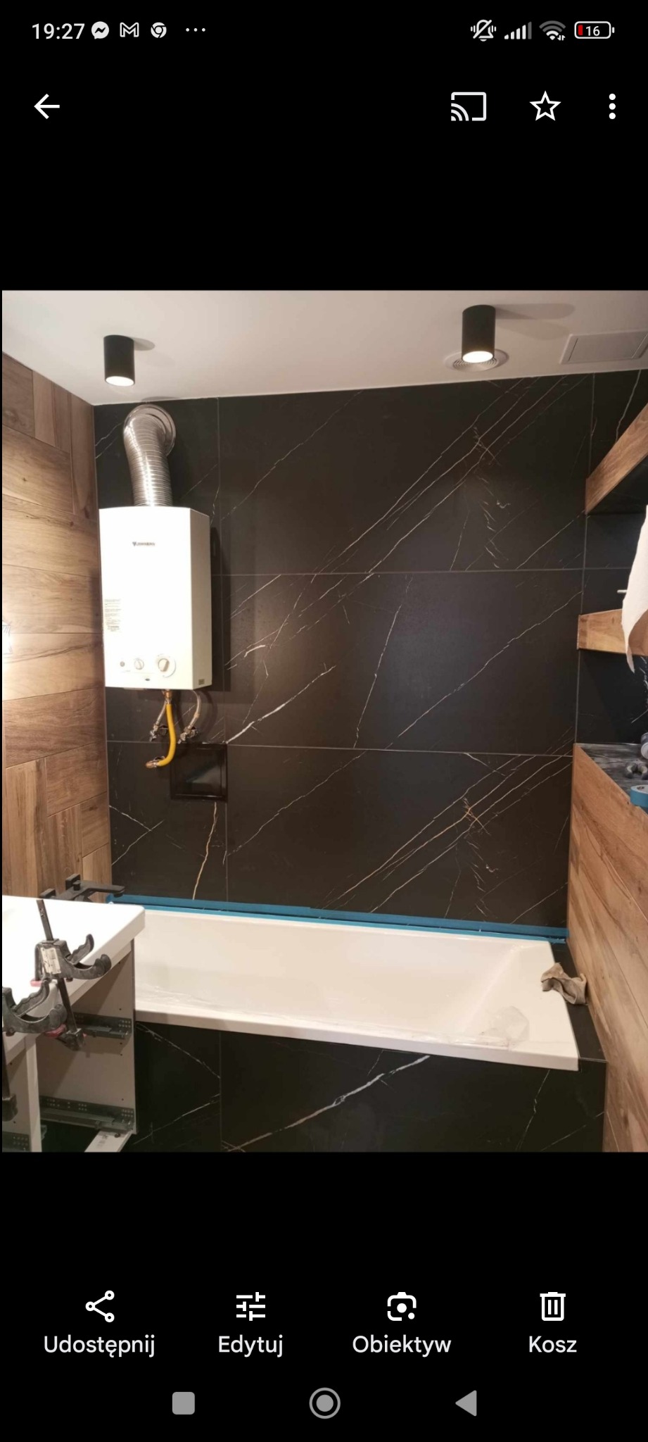 Nowoczesna łazienka z czarnymi płytkami imitującymi marmur, drewnianymi panelami, białą wanną i podgrzewaczem wody Junkers na ścianie.