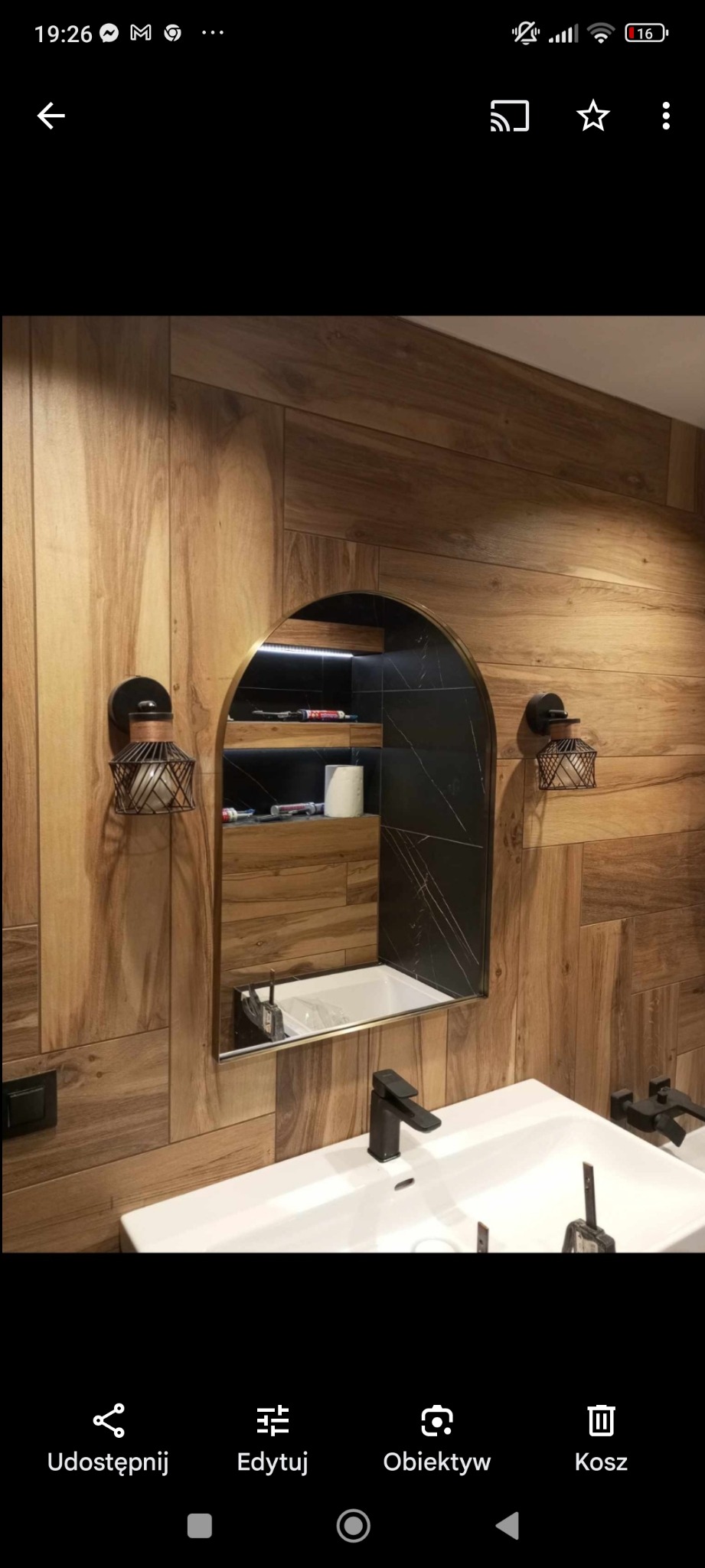 Nowoczesna łazienka z drewnopodobnymi płytkami na ścianach, białą umywalką z czarną armaturą, lustrem w kształcie łuku z czarnym obramowaniem i wnęką z oświetleniem LED, gdzie widoczne są akcesoria...