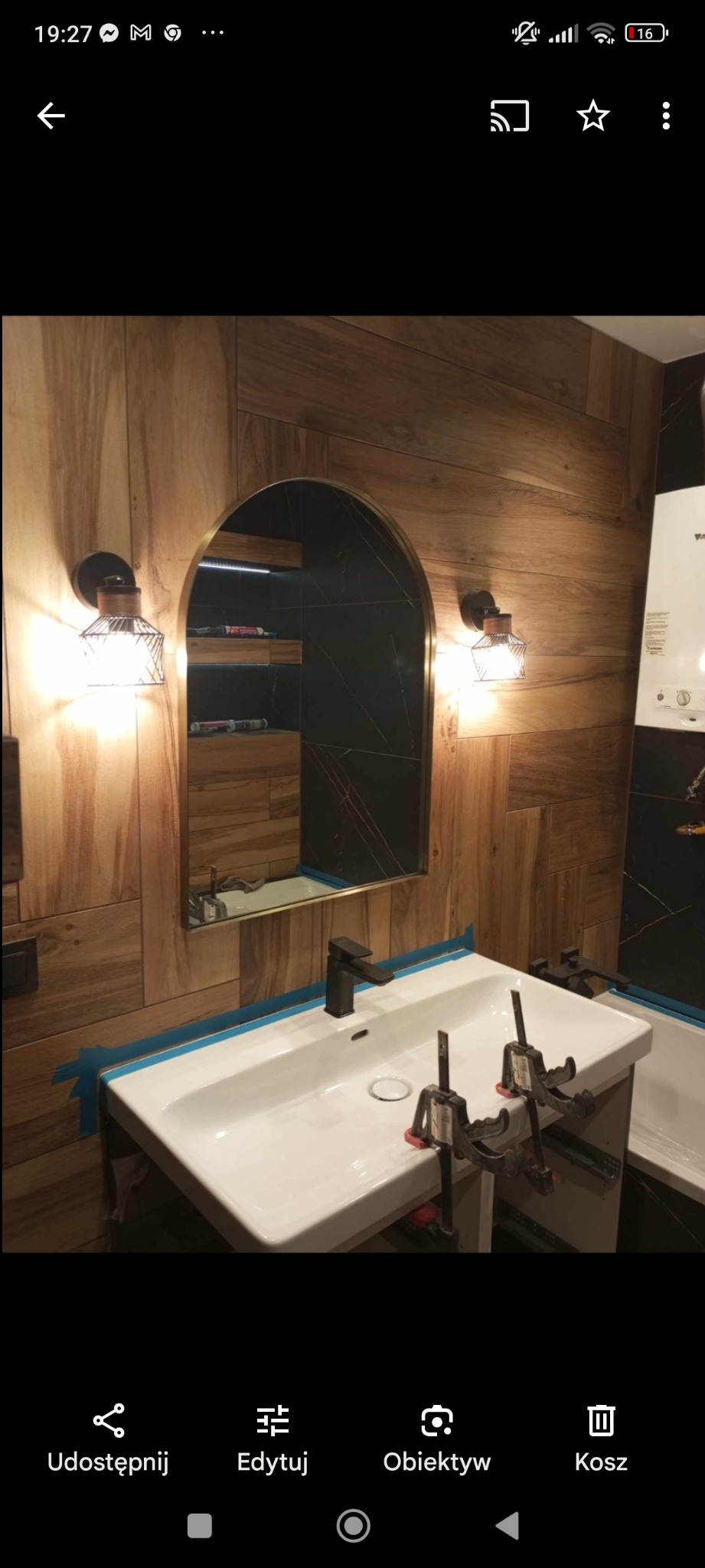 Montaż umywalki w łazience z drewnopodobnymi płytkami na ścianie i lustrem w kształcie łuku. Umywalka zabezpieczona taśmą malarską, widoczne klamry montażowe.