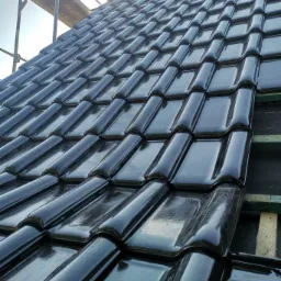 Błyszczące, czarne dachówki ceramiczne, ułożone na dachu z widoczną konstrukcją drewnianą i fragmentem rusztowania w tle, uchwycone pod kątem w słoneczny dzień.