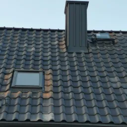 Ciemna dachówka ceramiczna z oknami dachowymi i metalowym kominem, widoczne ślady zabrudzeń i patyny na powierzchni dachu.