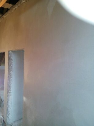 Świeżo nałożona, jasnoszara warstwa na ścianie wewnętrznej, widoczne nierówności i ślady po narzędziach, przygotowanie do dalszych prac wykończeniowych, sufit z widocznymi elementami konstrukcyjnymi.