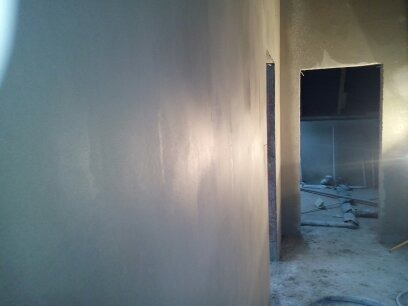 Świeżo nałożona, jasnoszara warstwa na ścianie, widoczne delikatne smugi po narzędziu, zarys futryny drzwiowej i widok na wnętrze pomieszczenia w trakcie remontu.