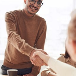 Legalizacja GC Biuro Obsługi Cudzoziemców - Uśmiechnięty mężczyzna w okularach w brązowym swetrze podaje dłoń kobiecie na jasnym drewnianym biurku z notatnikiem, długopisem i kubkami.