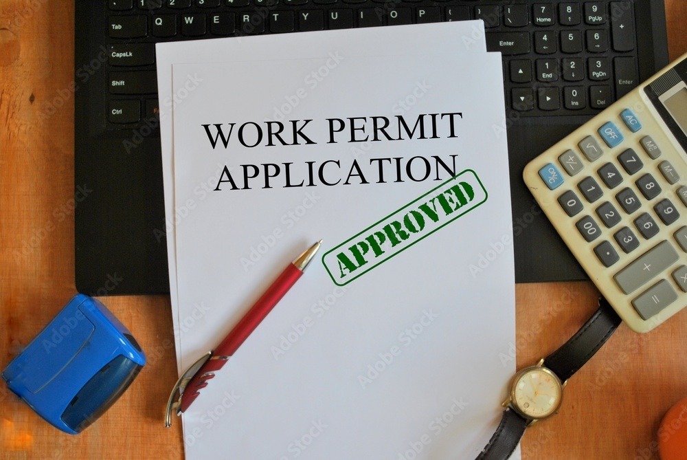 Dokument z napisem 'Work Permit Application' i zielonym stemplem 'APPROVED' leży na biurku obok kalkulatora, długopisu i pieczątki, widoczna klawiatura laptopa w tle.