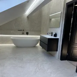 Elegancka łazienka na poddaszu ze skośnym sufitem, płytki imitujące marmur, wolnostojąca wanna, czarna szafka pod umywalką i czarne drzwi z ozdobnymi panelami.