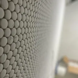 Ściana wyłożona mozaiką z białych, okrągłych płytek z delikatnym marmurkowym wzorem, ułożonych w regularny wzór, z rozmytym tłem wnętrza łazienki.