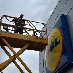 Szop Czyściec Karolina Markiewicz - Pracownik na podnośniku koszowym, na tle szarej elewacji z logo. Osoba w kombinezonie, widok z dołu na kosz i część elewacji budynku.