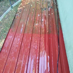 Częściowo umyta, czerwona blacha trapezowa na dachu domu. Widoczna granica czystości i resztki liści. W tle zieleń i fragment ściany budynku.