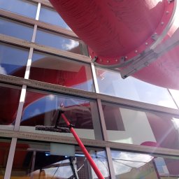 Szop Czyściec Karolina Markiewicz - Mycie okien na wysokości z użyciem teleskopowego kija, widoczna czerwona konstrukcja rurowa i refleksy nieba w czyszczonych oknach.