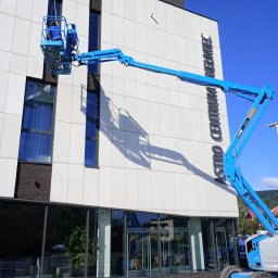 Szop Czyściec Karolina Markiewicz - Pracownik w niebieskim podnośniku koszowym myje okna nowoczesnego budynku Astro Centrum Chełmiec. Widoczne duże okna i biała elewacja budynku.