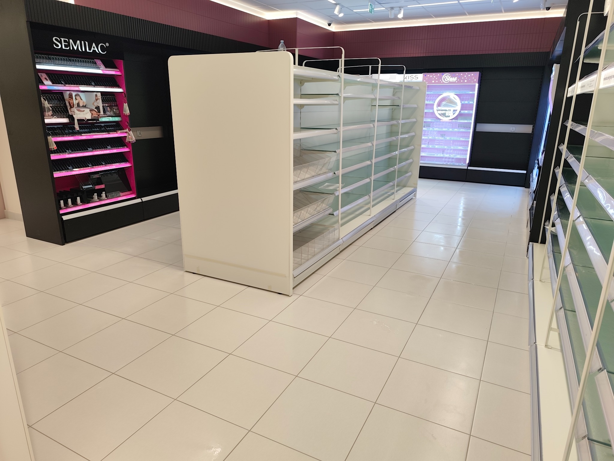 Półki sklepowe, białe, z przezroczystymi szklanymi półkami, w jasnym wnętrzu sklepu. Widoczne regały z kosmetykami w tle. Perspektywa z dołu.