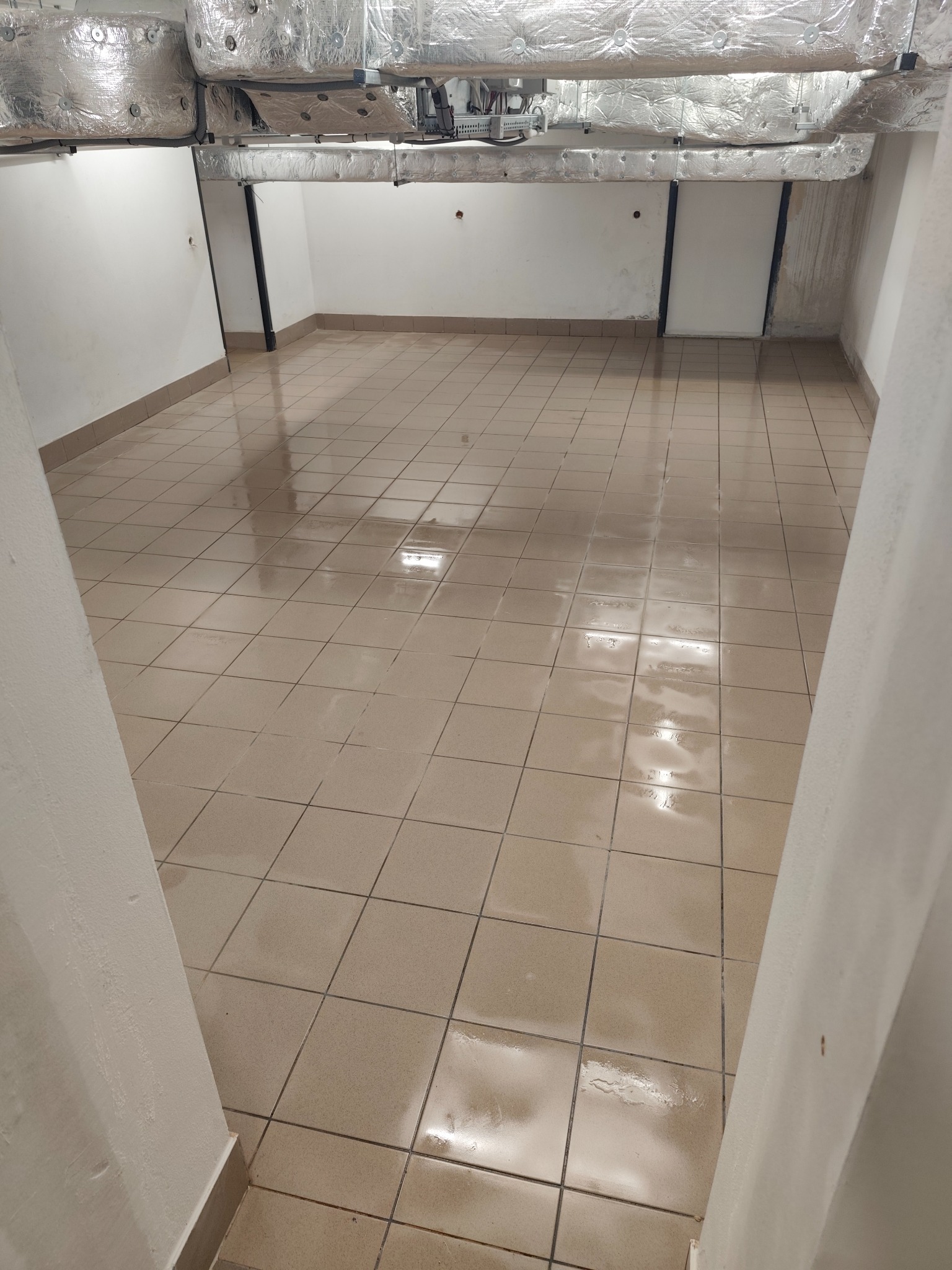 Wykafelkowane pomieszczenie z widocznymi instalacjami pod sufitem. Podłoga po umyciu, widać refleksy wody. Ściany białe, w tle widoczne drzwi.