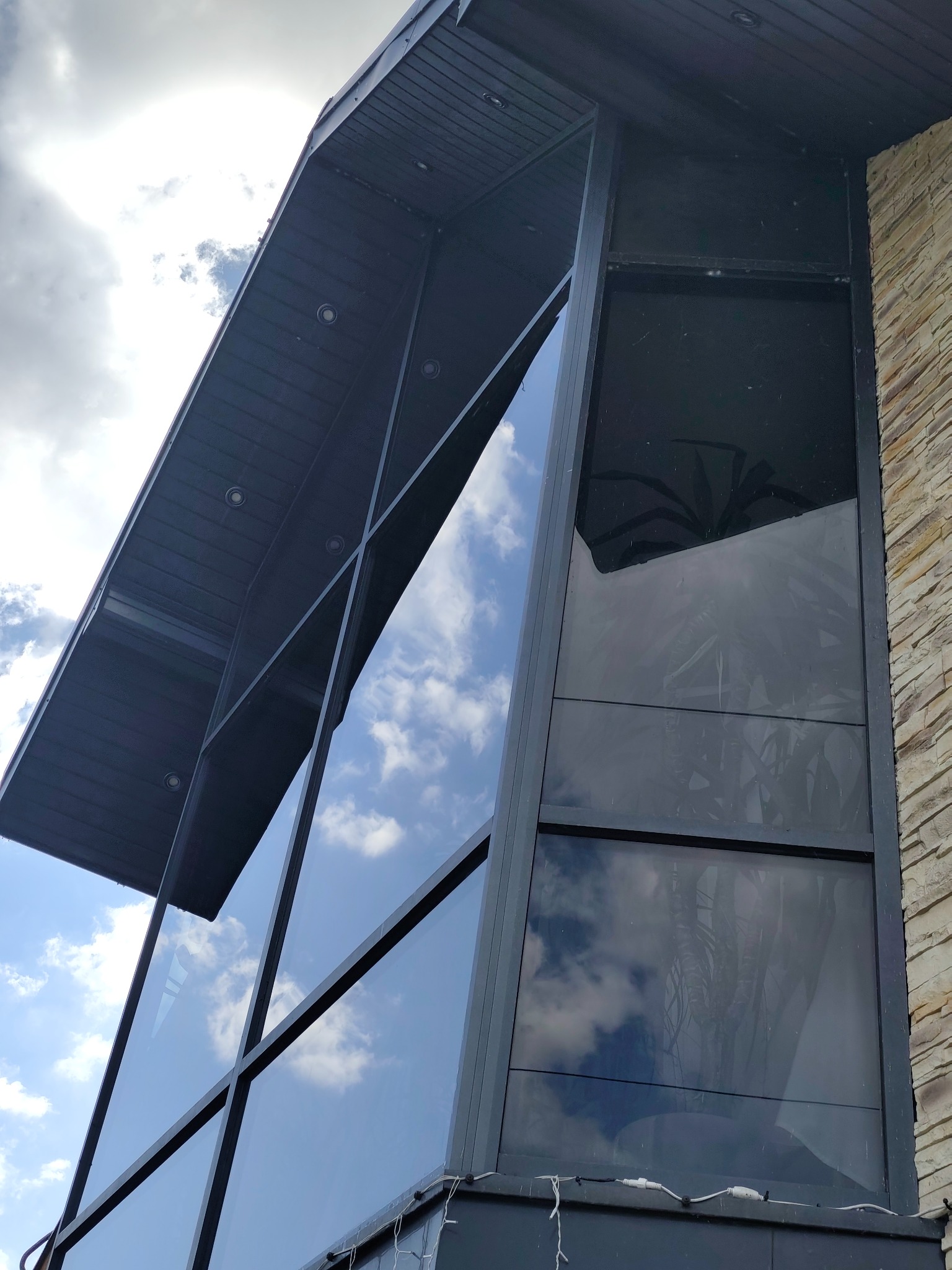 Nowoczesna fasada budynku z dużymi, czystymi oknami, w których odbija się błękitne niebo z chmurami. Widoczna kamienna elewacja i fragment dachu.