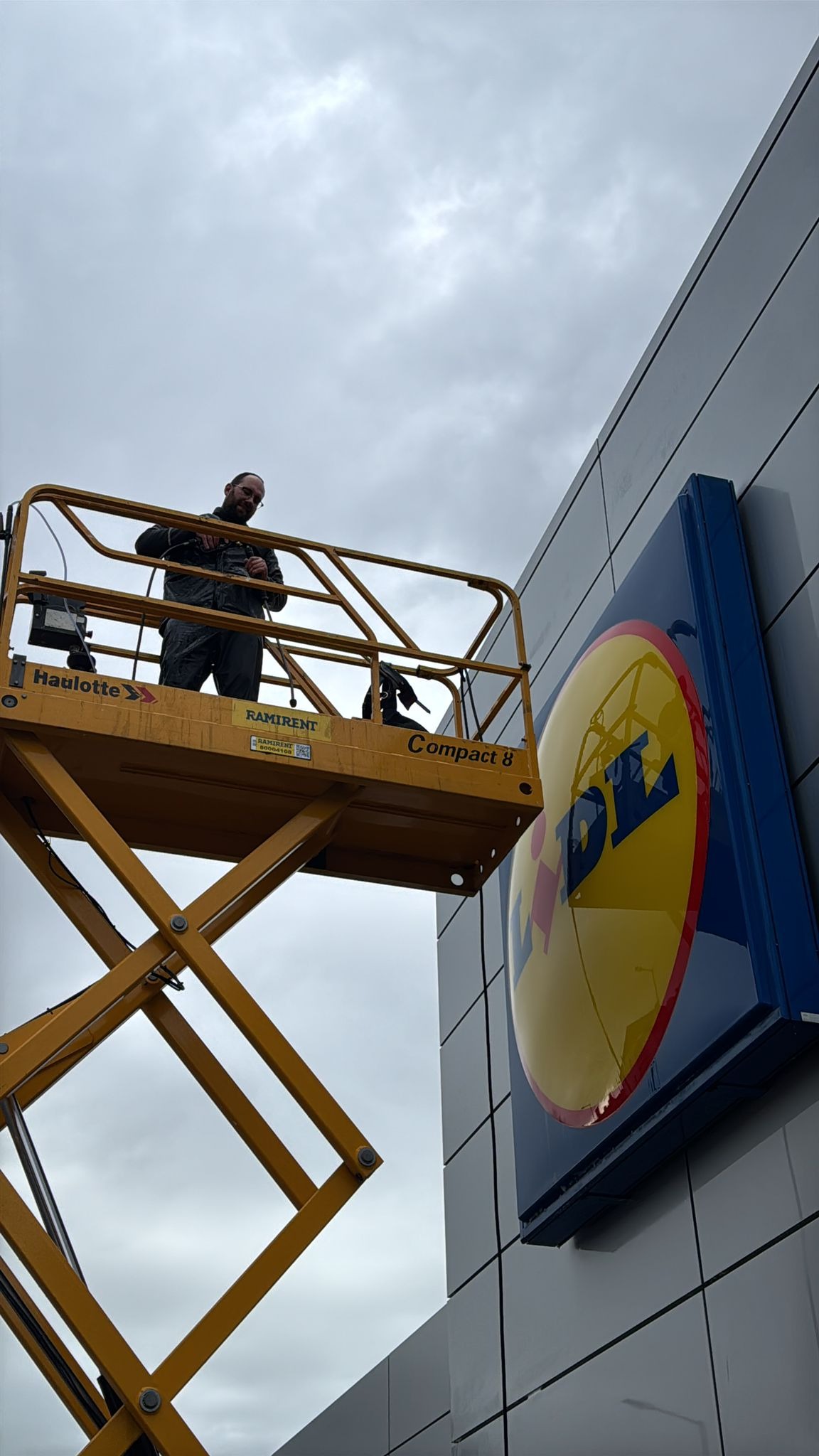 Pracownik na podnośniku koszowym, na tle szarej elewacji z logo. Osoba w kombinezonie, widok z dołu na kosz i część elewacji budynku.