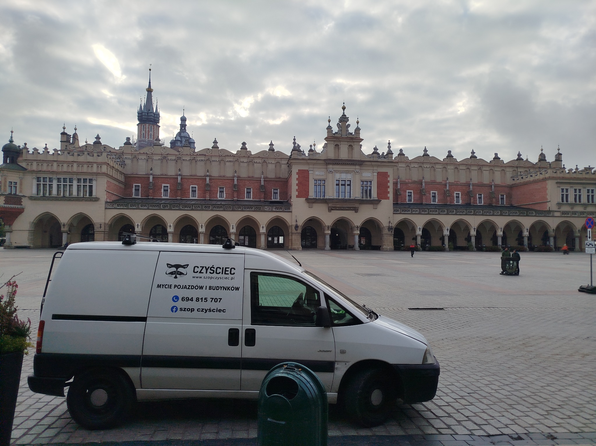 Usługa mycia pojazdów i budynków: biały van firmy 'Czyściec' na tle Sukiennic w Krakowie. Napis na samochodzie: 'Mycie pojazdów i budynków'.