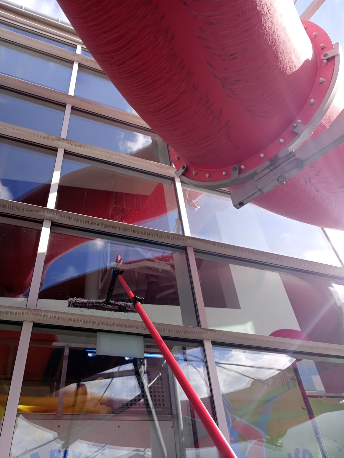Mycie okien na wysokości z użyciem teleskopowego kija, widoczna czerwona konstrukcja rurowa i refleksy nieba w czyszczonych oknach.