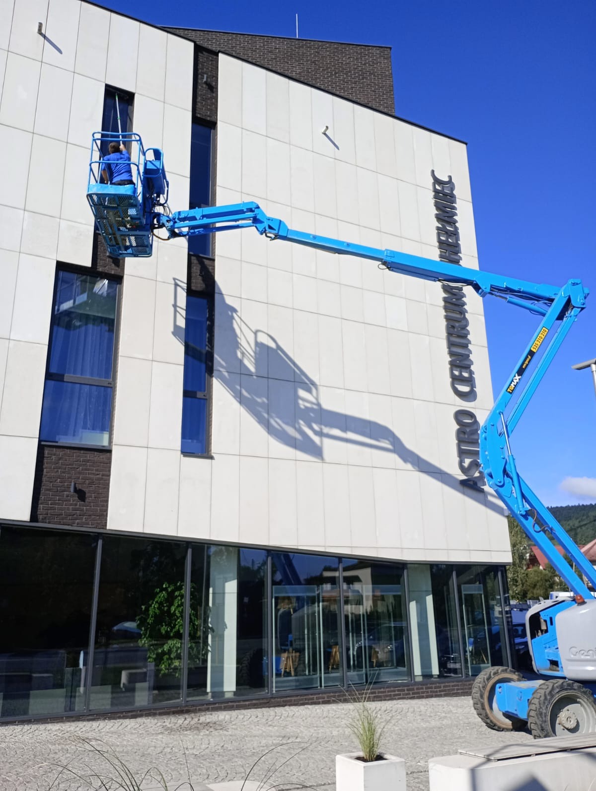 Pracownik w niebieskim podnośniku koszowym myje okna nowoczesnego budynku Astro Centrum Chełmiec. Widoczne duże okna i biała elewacja budynku.