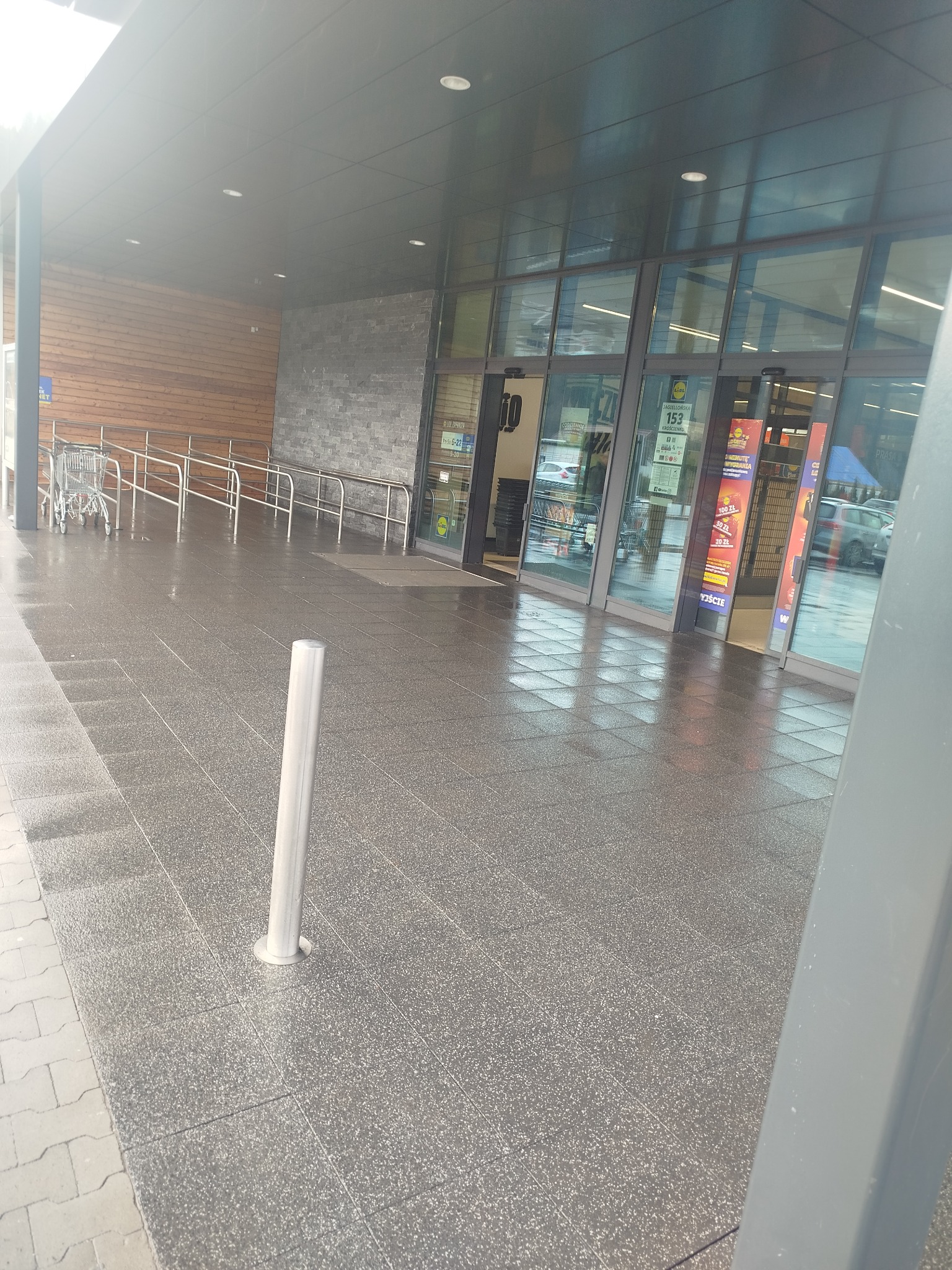 Błyszcząca, mokra kostka brukowa przed wejściem do sklepu, z widocznymi wózkami i rampą dla niepełnosprawnych. Słupki zabezpieczające. Widoczne reklamy na szybach.