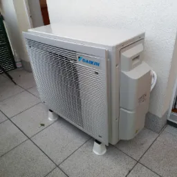 Zewnętrzna jednostka klimatyzacji Daikin zamontowana na balkonie, widoczna z niskiej perspektywy, z detalami wykończenia balkonu i fragmentem krzesła w tle.