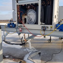 SYSCON - Agregat chłodniczy na dachu budynku komercyjnego z podłączonymi pompami próżniowymi i butlą z gazem technicznym, widoczny manometr i przewody, w tle zimowy krajobraz.