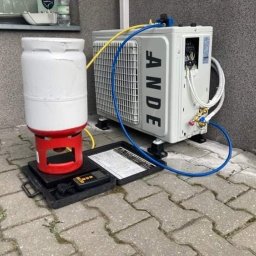 SYSCON - Serwis klimatyzacji: biała jednostka zewnętrzna klimatyzacji marki ANDE podłączona przewodami do butli z czynnikiem chłodniczym, umieszczonej na wadze elektronicznej.