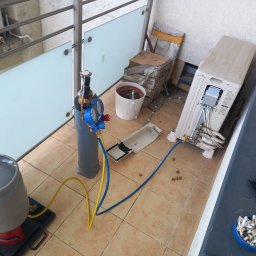 SYSCON - Sprzęt do napełniania klimatyzacji na balkonie, butla z manometrami, węże żółty i niebieski, rozmontowana jednostka zewnętrzna, popielniczka z niedopałkami.