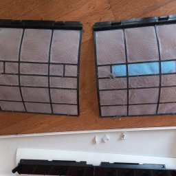 SYSCON - Dwa wyjęte, zakurzone filtry klimatyzacji z czarną ramką, jeden z niebieskim wkładem, leżące obok otwartej, białej obudowy klimatyzatora na drewnianej podłodze.