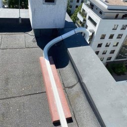 SYSCON - Biała, karbowana rura wentylacyjna podłączona do komina na dachu budynku mieszkalnego, zabezpieczona cegłą, z widocznymi budynkami mieszkalnymi w tle.